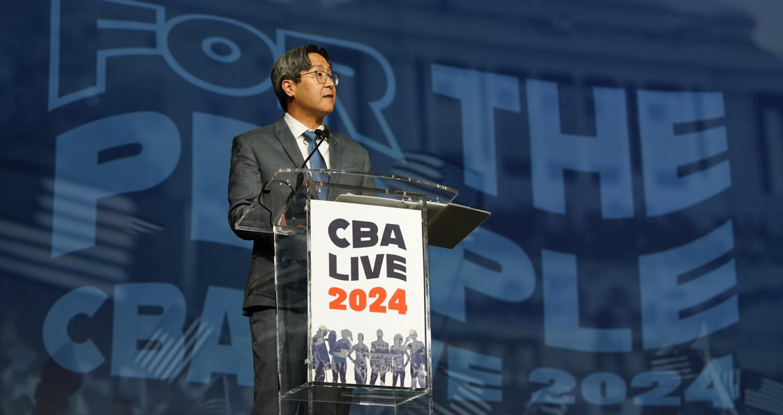 Acting Comptroller Hsu stands at a clear podium with a 'CBA Live 2024' sign against a backdrop reading For the People.