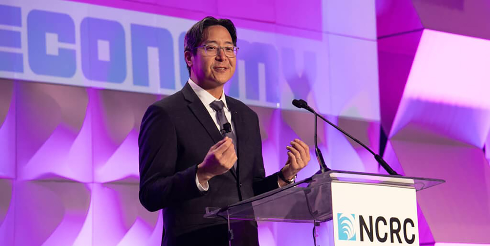 Acting Comptroller Hsu delivers remarks at a clear podium with an NCRC sign, set against a pink and purple-lit backdrop featuring the word 'Economy'