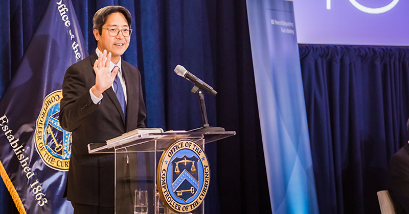 Image of Acting Comptroller of the Currency Michael J. Hsu has his hand up while speaking from a clear lectern with an Office of the Comptroller of the Currency logo. He is standing in front of an Office of the Comptroller of the Currency flag.