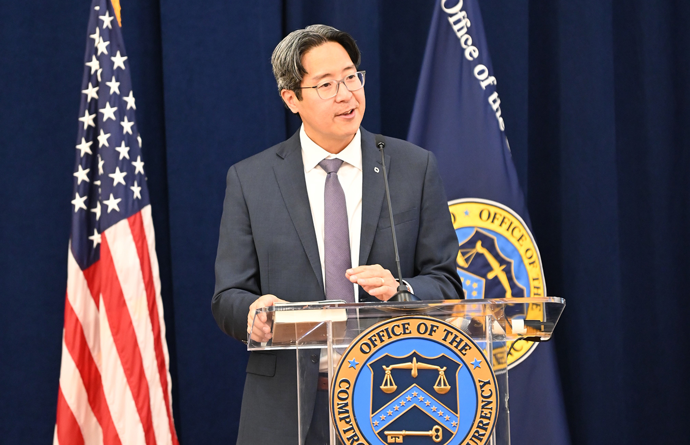 Acting Comptroller Hsu speaks at a clear podium adorned with the Office of the Comptroller of the Currency seal, flanked by the U.S. flag and the OCC flag against a dark blue backdrop.